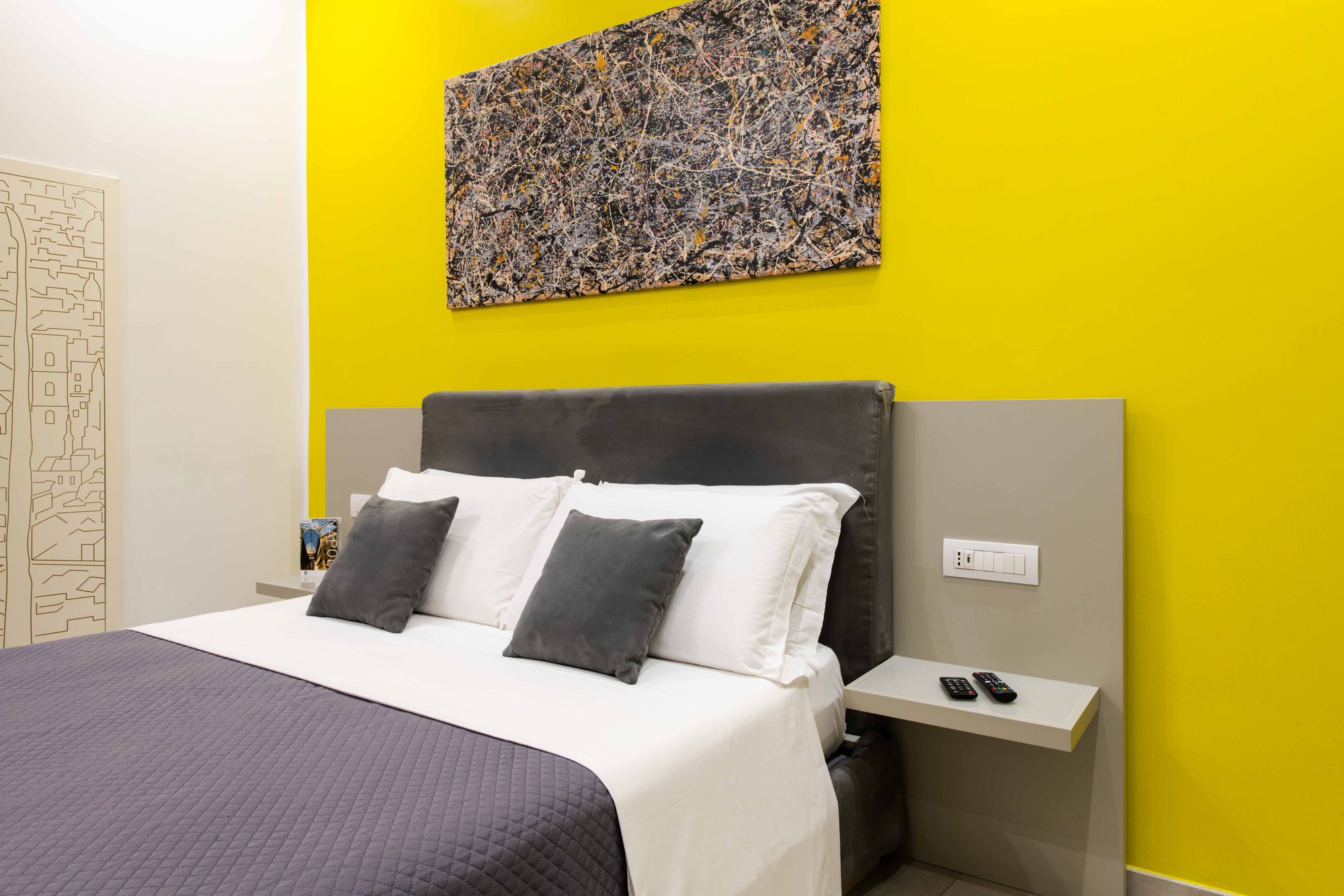 Dettaglio testiera e cuscini del letto matrimoniale con telecomandi sul comodino sospeso, parete gialla con quadro astratto - Toledo16 Luxury Suites Napoli