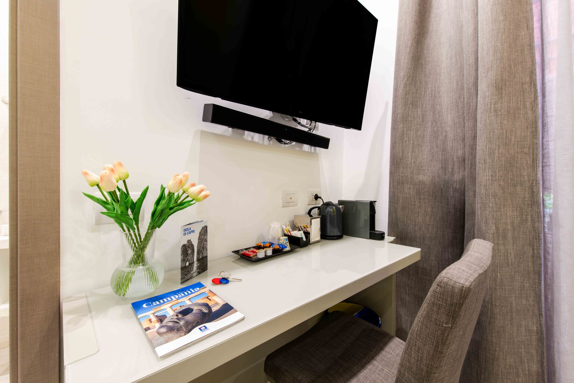 Scrivania con vaso di tulipani, guide turistiche di Capri e Campania, chiavi della camera, vassoio di snack, bollitore e macchina da caffè sotto TV a parete - Toledo16 Luxury Suites Napoli