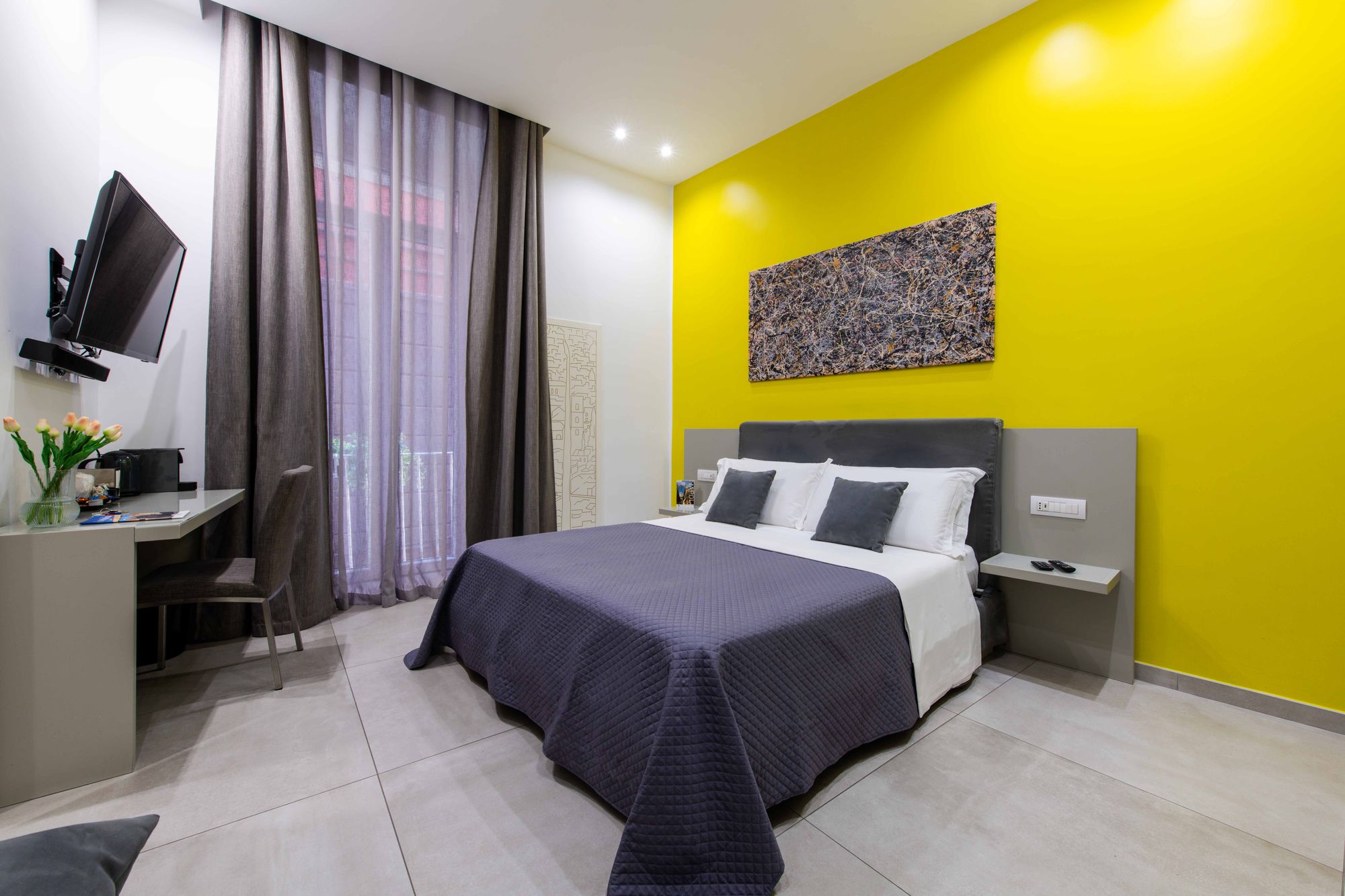 Camera con parete gialla, TV a schermo piatto a muro, scrivania con macchina da caffè e vaso di tulipani, tende grigie sulla porta finestra affacciata su Via Toledo - Toledo16 Luxury Suites Napoli