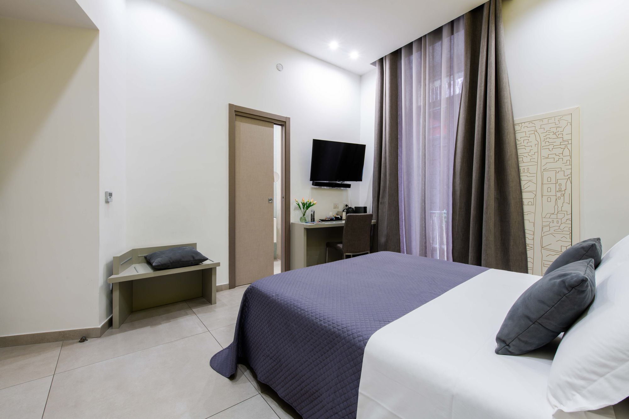 Vista d'insieme della camera con panca imbottita, porta del bagno aperta, TV a parete e pannello decorativo con skyline di Napoli, B&B Napoli centro - Toledo16 Luxury Suites Napoli