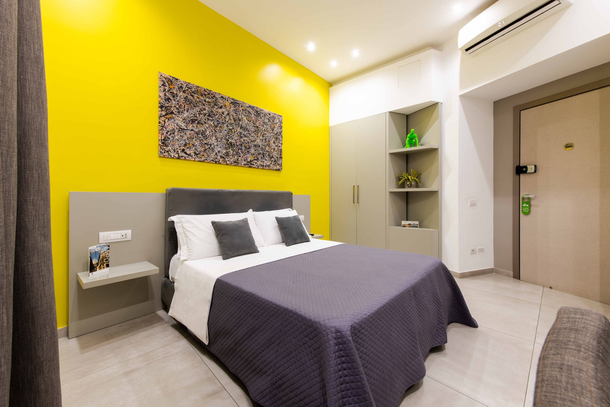 Camera con armadio e libreria d'angolo con statuetta verde e piantina decorativa, porta d'ingresso con serratura magnetica, testiera grigia su parete gialla - Toledo16 Luxury Suites Napoli