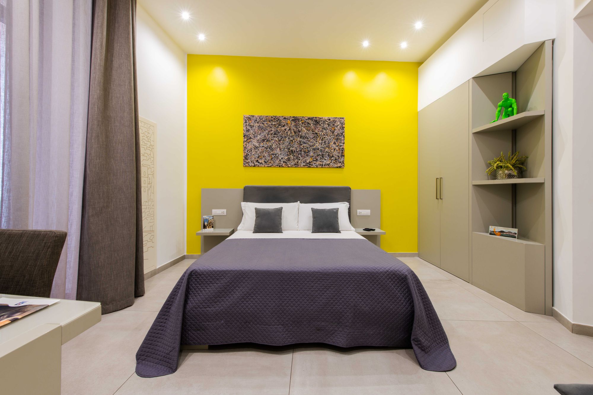 Letto matrimoniale visto frontalmente con testiera grigia, quadro astratto su parete gialla, armadio e mensole d'angolo con statuetta verde, camera B&B Napoli - Toledo16 Luxury Suites Napoli