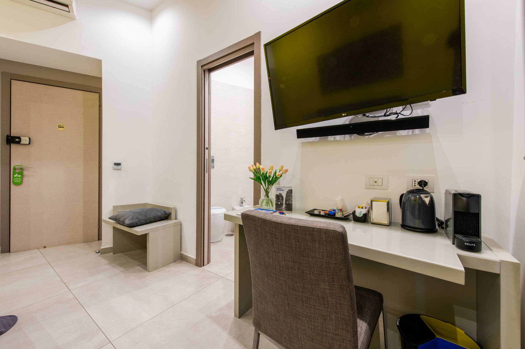 Scrivania con TV a parete, bollitore, macchina da caffè Krups e vaso di tulipani, panca con cuscino grigio e porta d'ingresso sullo sfondo - Toledo16 Luxury Suites Napoli