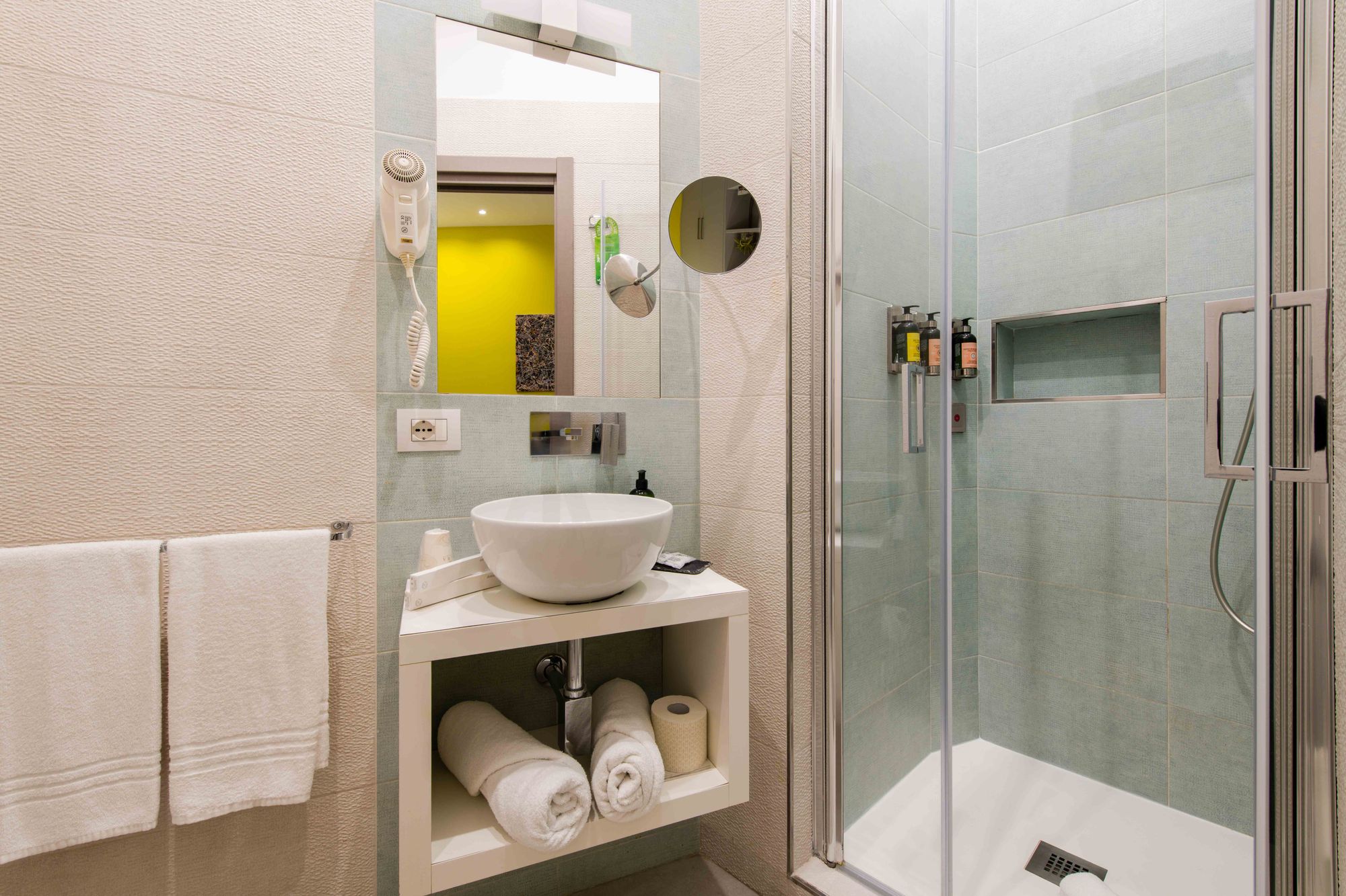Bagno con lavabo bianco a bacinella, asciugacapelli a parete, specchio con luce e specchietto ingranditore, asciugamani arrotolati e box doccia in vetro con piastrelle azzurre - Toledo16 Luxury Suites Napoli