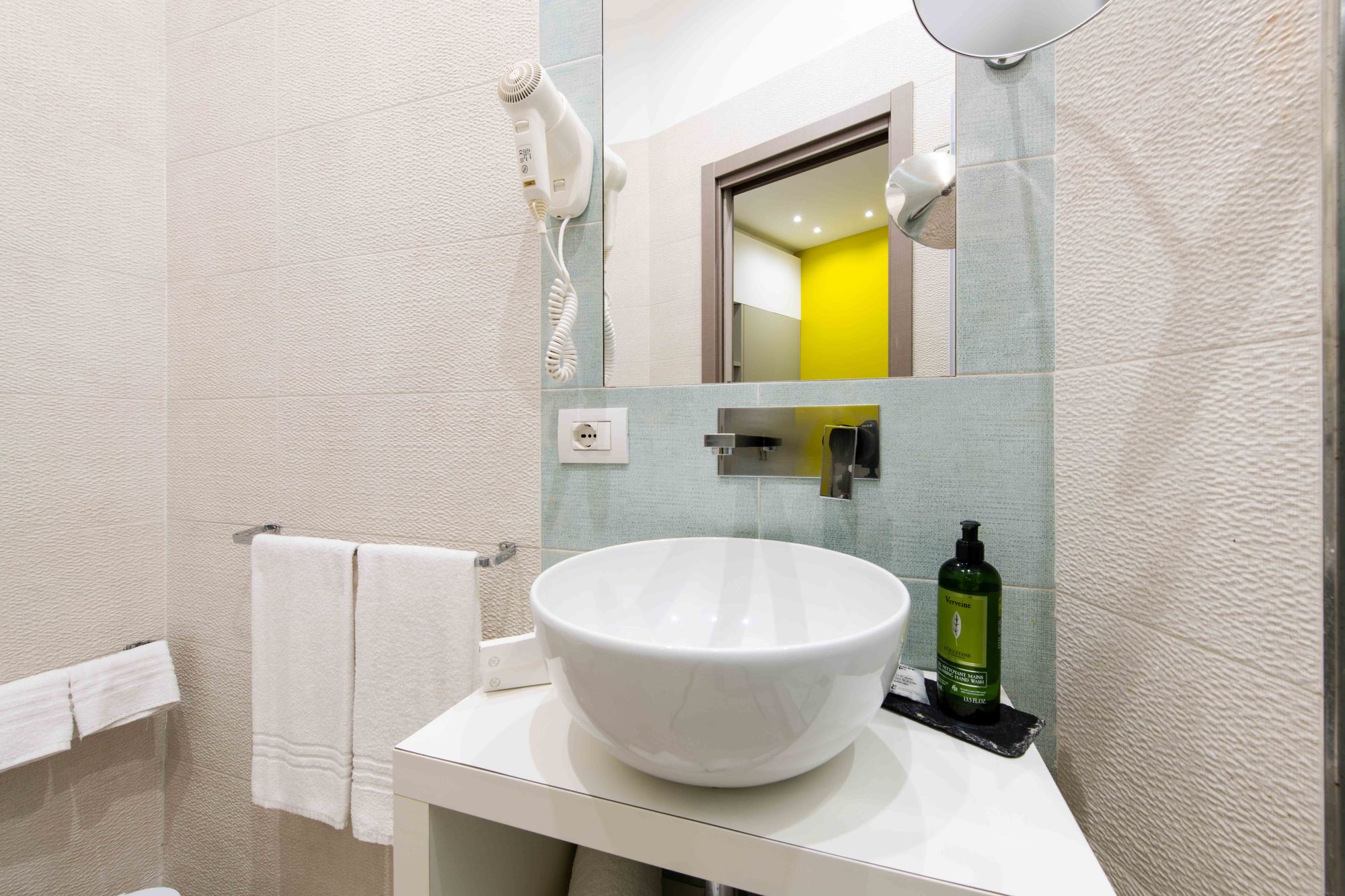Dettaglio lavabo a bacinella bianca con rubinetteria cromata a parete, dispenser di sapone L'Occitane, specchio e asciugacapelli - Toledo16 Luxury Suites Napoli