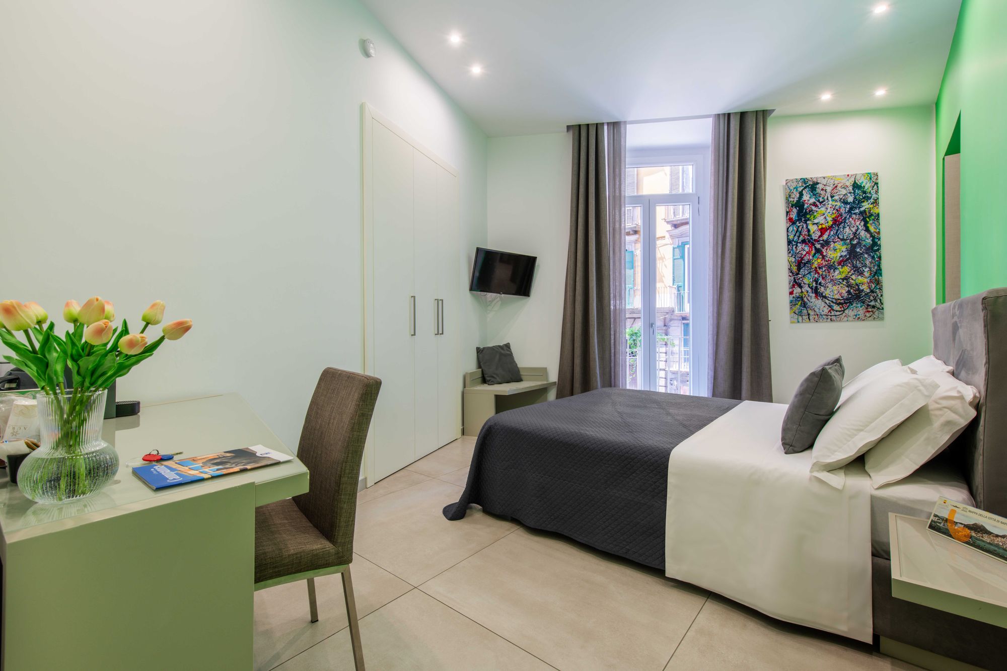 Camera con scrivania verde salvia, sedia imbottita marrone e vaso di tulipani, letto con copriletto grigio antracite e finestra con balconcino su Via Toledo - Toledo16 Luxury Suites Napoli