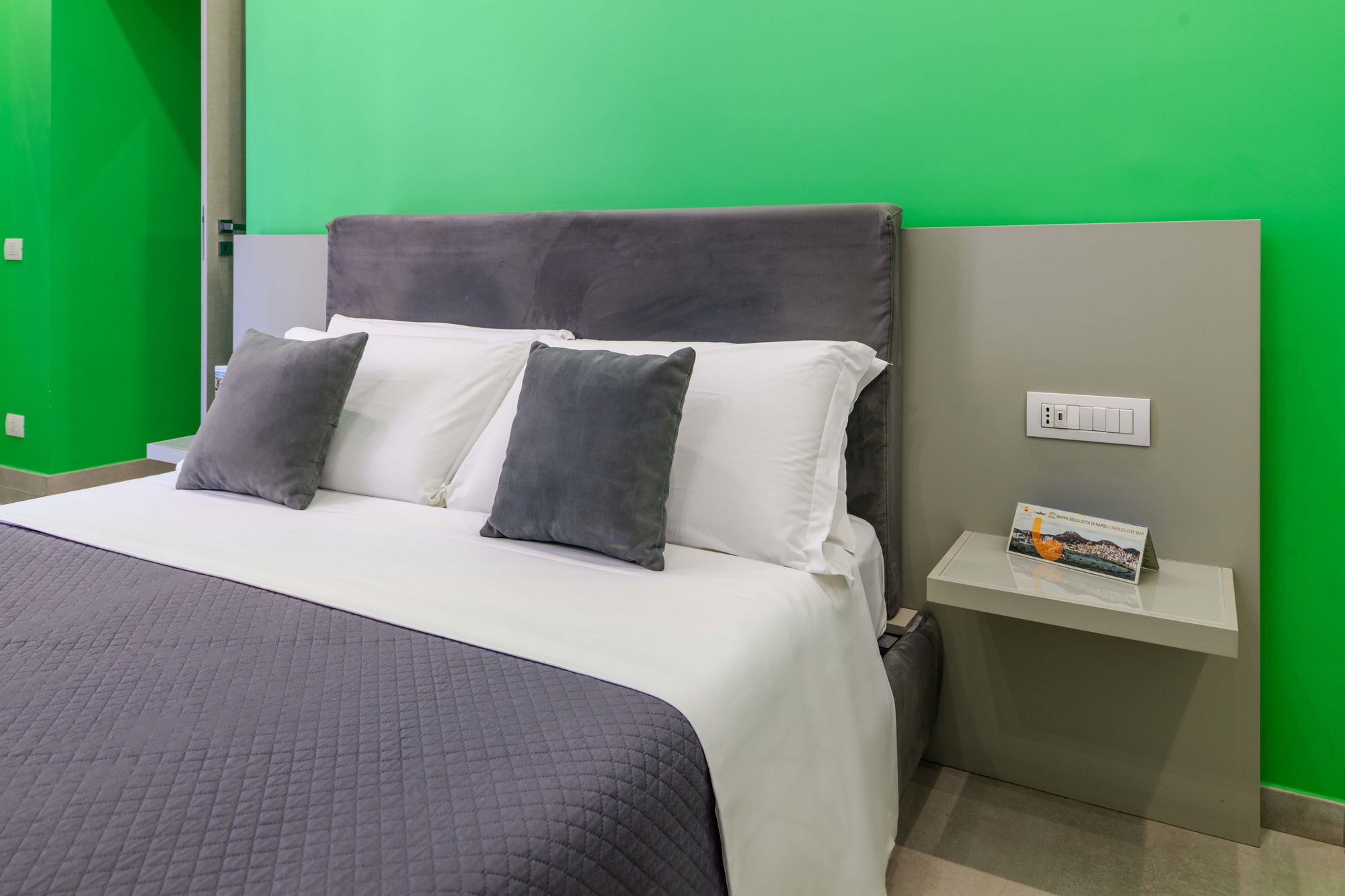Dettaglio comodino sospeso con guida di Napoli, testiera in velluto grigio con cuscini bianchi e grigi su parete verde smeraldo - Toledo16 Luxury Suites Napoli