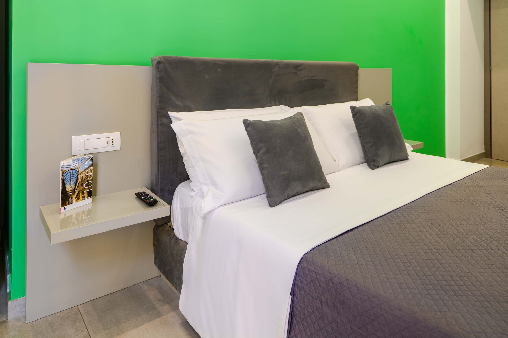 Comodino sospeso con guida di Napoli e telecomando, vista ravvicinata sui cuscini grigi e bianchi del letto - Toledo16 Luxury Suites Napoli