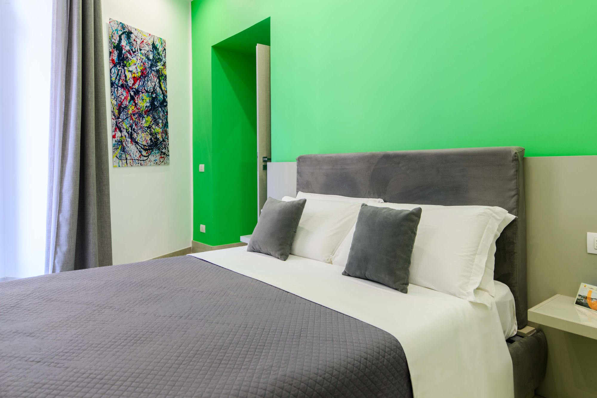 Letto visto dal corridoio, testiera grigia e cuscini su parete verde smeraldo con quadro astratto colorato, porta del bagno sullo sfondo - Toledo16 Luxury Suites Napoli