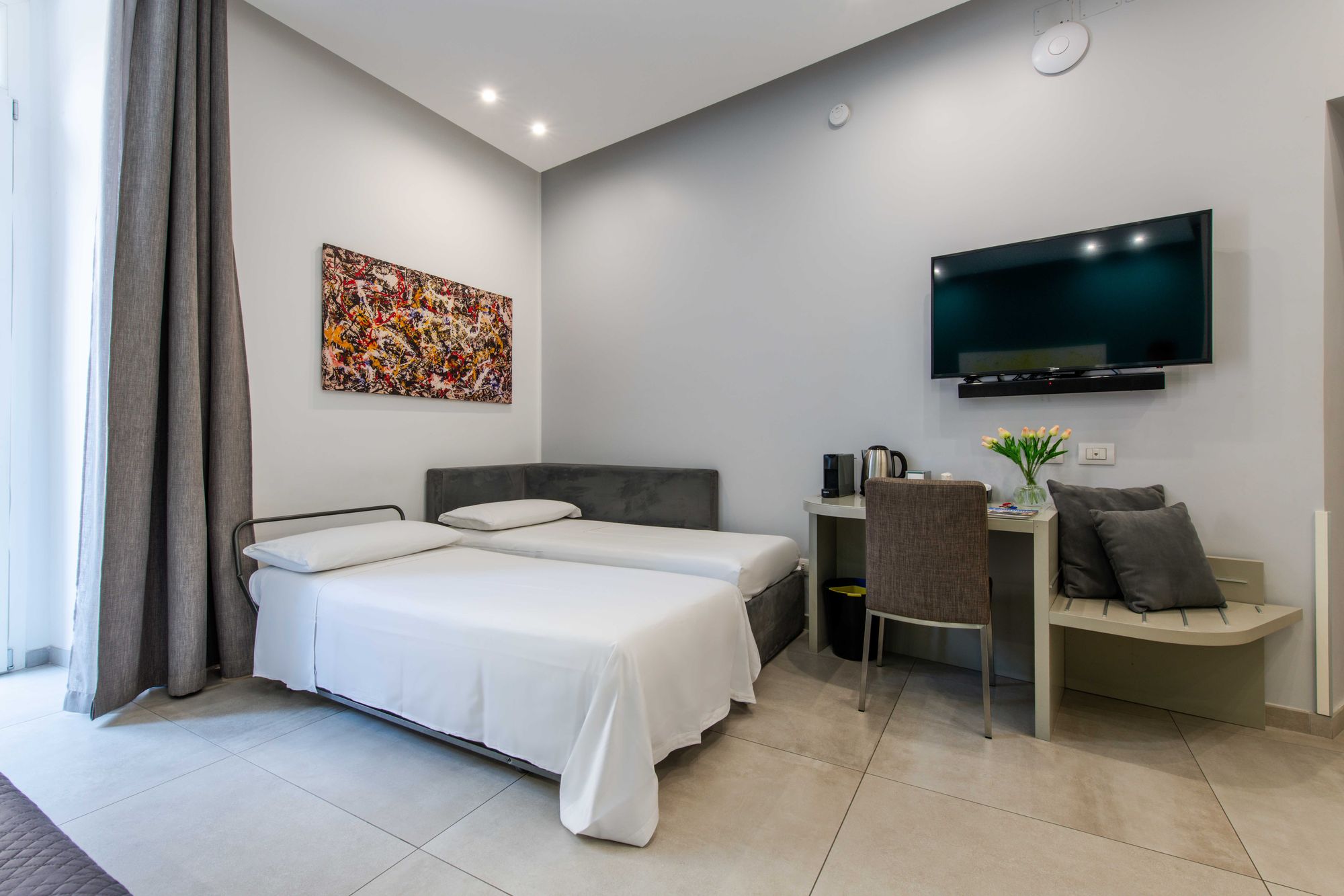 Letto aggiuntivo con lenzuola bianche accanto al divano letto grigio, televisore a parete e scrivania della suite - Toledo16 Luxury Suites Napoli