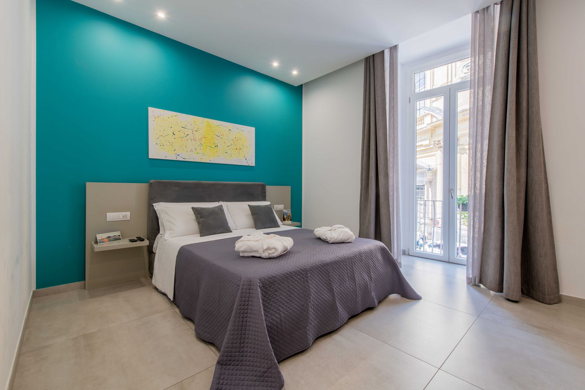 Camera con parete turchese, testata del letto in velluto grigio, quadro astratto giallo e porta finestra con vista sulla strada - Toledo16 Luxury Suites Napoli