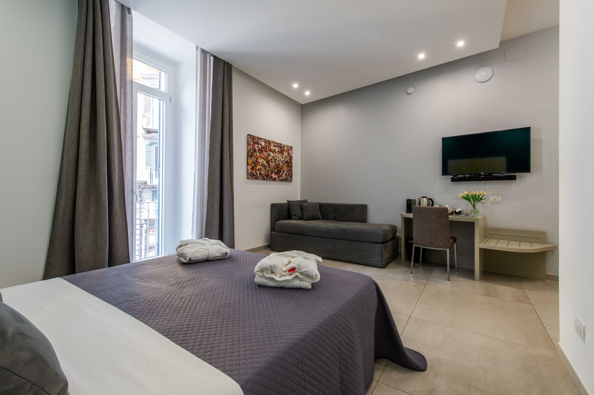 Zona soggiorno della suite con divano grigio, televisore a parete, quadro astratto colorato e scrivania con bollitore e fiori - Toledo16 Luxury Suites Napoli