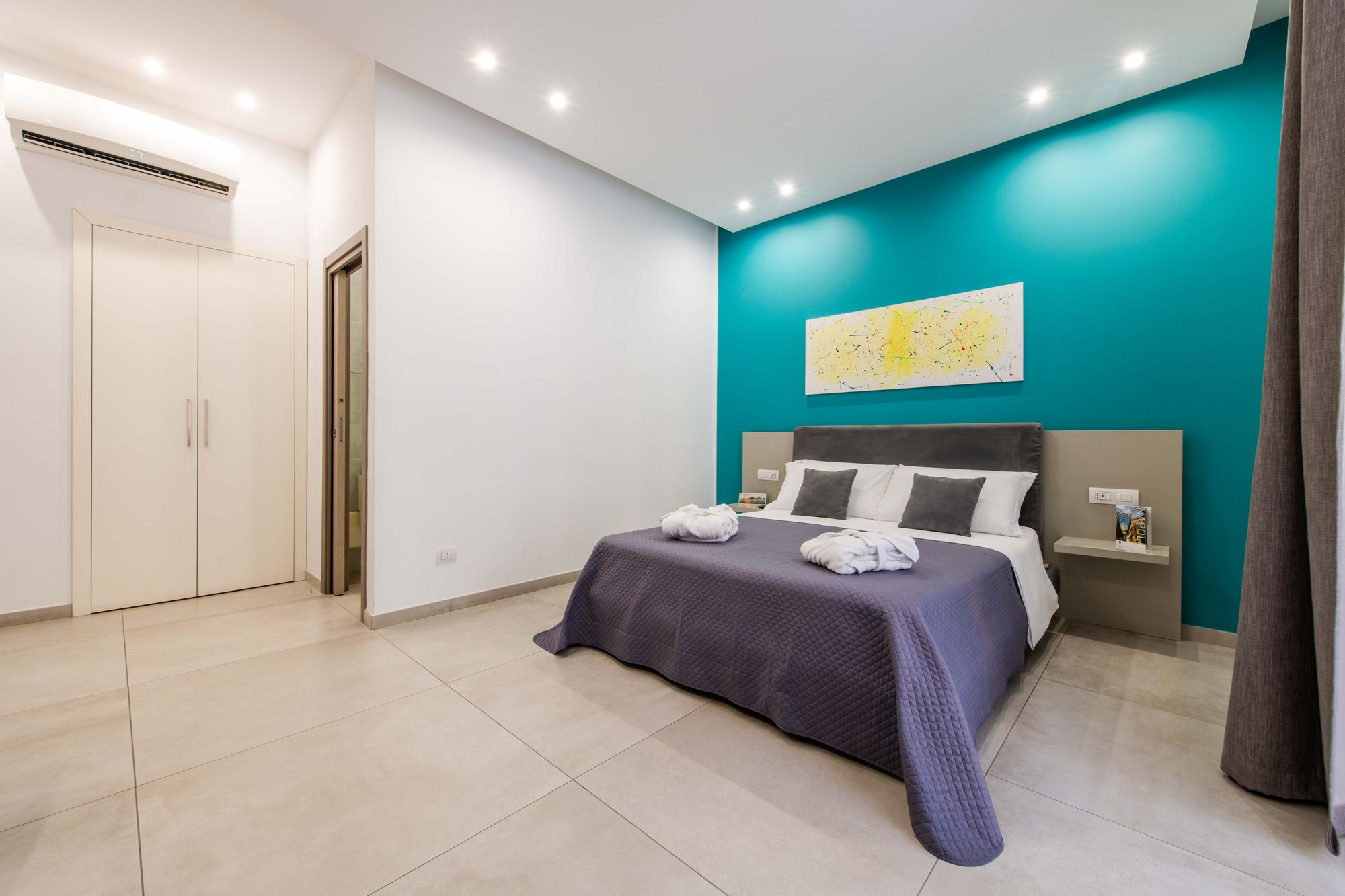 Vista dell'armadio guardaroba e della camera con parete turchese, letto matrimoniale e accappatoi piegati sul copriletto - Toledo16 Luxury Suites Napoli