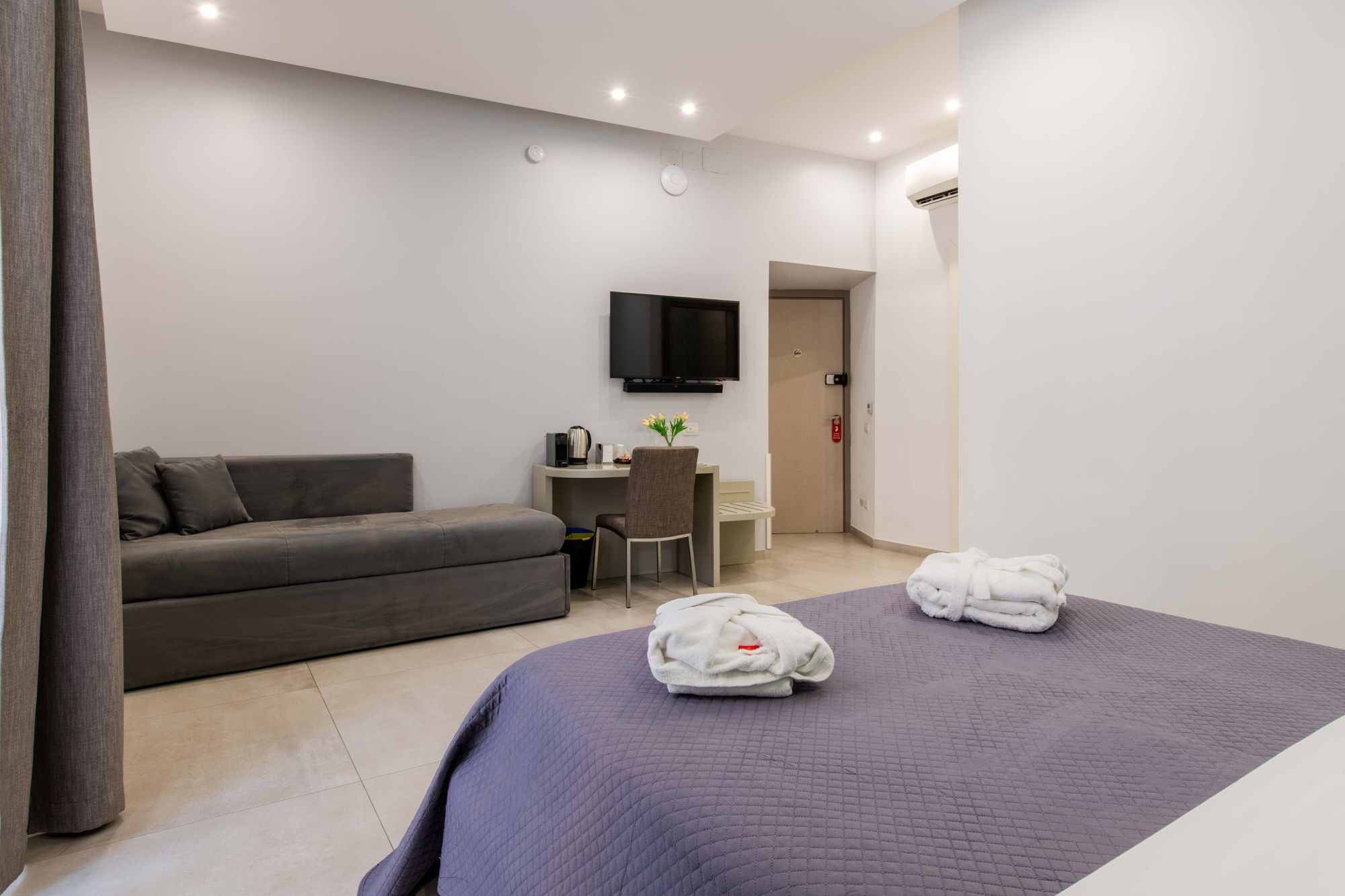 Angolo soggiorno con divano grigio, televisore a parete, scrivania e climatizzatore, con letto matrimoniale in primo piano - Toledo16 Luxury Suites Napoli