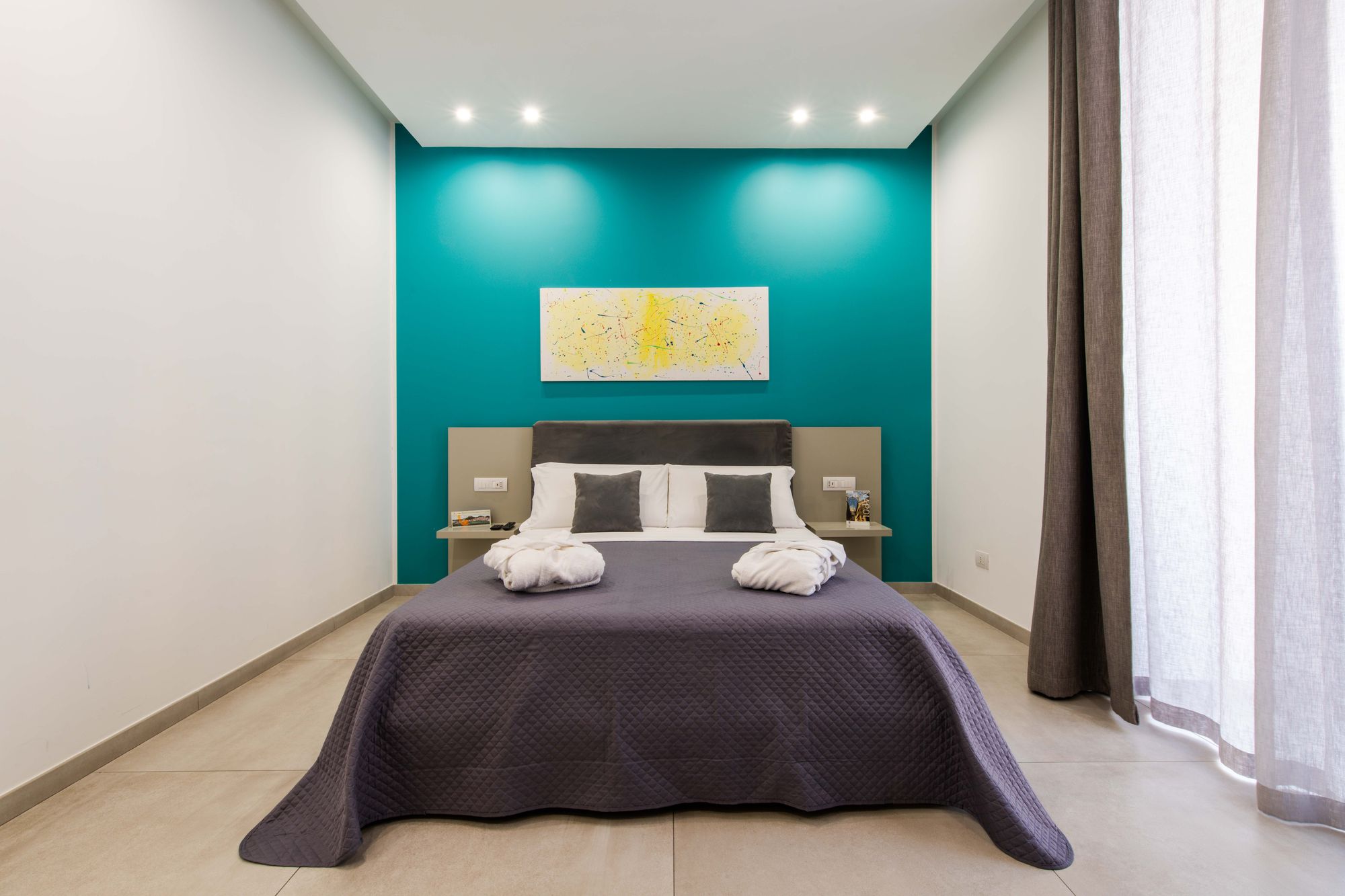 Letto matrimoniale con testata in velluto grigio, cuscini decorativi, quadro astratto giallo su parete turchese e accappatoi piegati sul copriletto - Toledo16 Luxury Suites Napoli