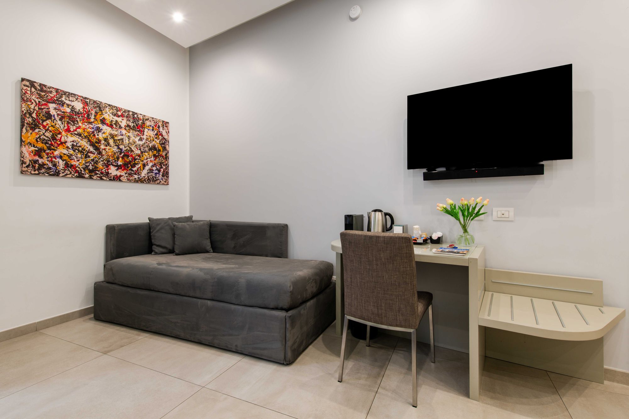 Zona soggiorno con divano letto grigio, quadro astratto colorato, televisore a parete e scrivania con set di benvenuto e fiori - Toledo16 Luxury Suites Napoli
