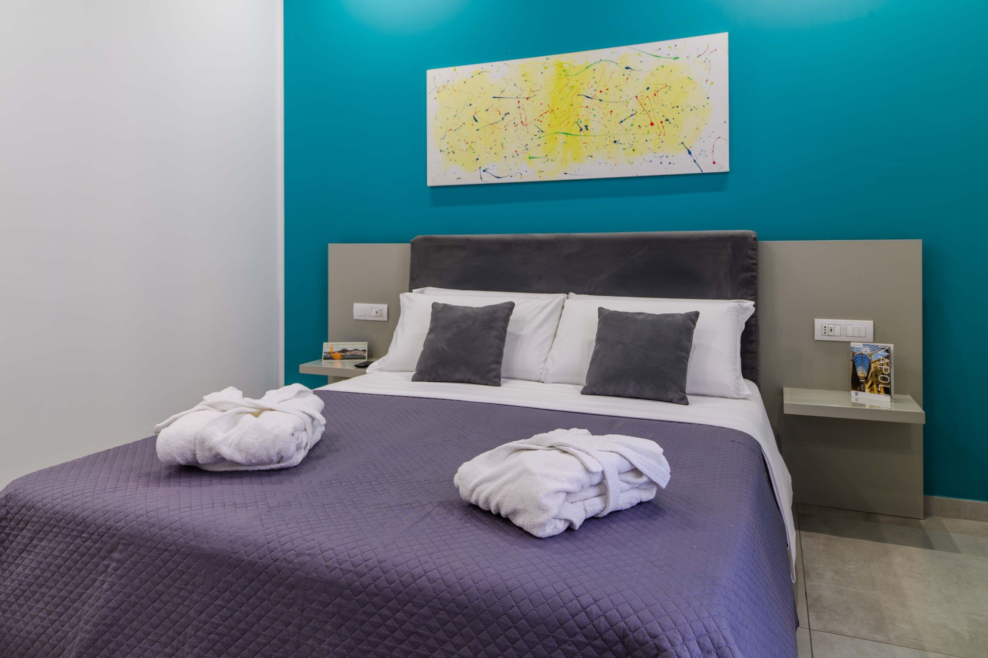 Letto matrimoniale con accappatoi piegati sul copriletto viola, cuscini grigi e quadro astratto giallo sulla parete turchese - Toledo16 Luxury Suites Napoli