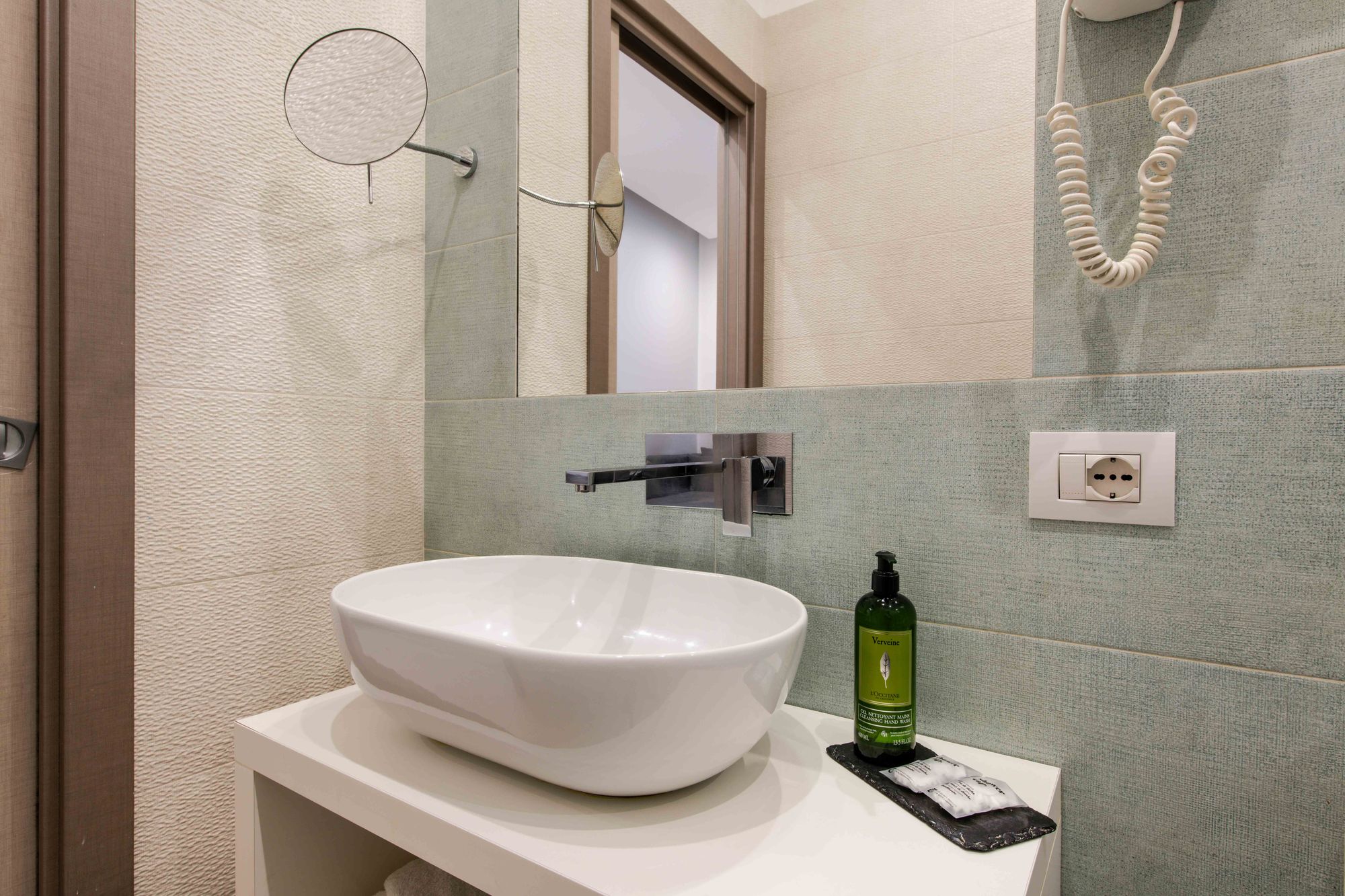 Dettaglio del lavabo del bagno con specchio, asciugacapelli a parete e prodotti da bagno L'Occitane - Toledo16 Luxury Suites Napoli