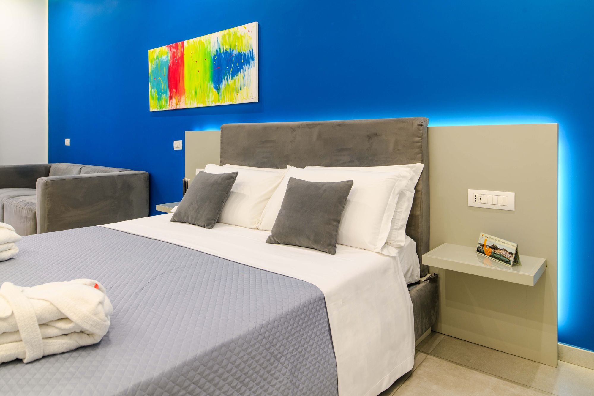 Divano grigio in velluto con tavolino in vetro e metallo, tende alla porta finestra del balcone, parete blu con quadro - Toledo16 Luxury Suites Napoli