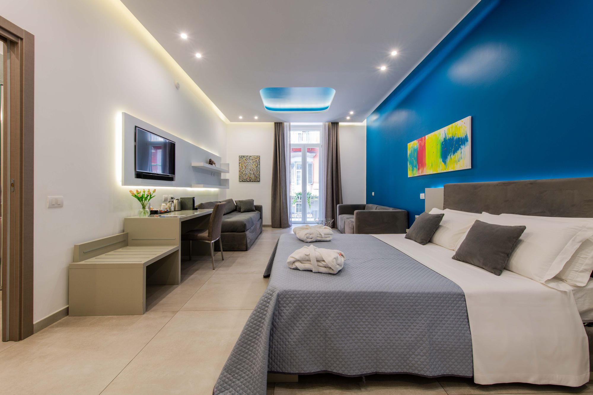 Suite con parete blu e quadro astratto multicolore, letto matrimoniale con plaid grigio e accappatoi, divano e scrivania - Toledo16 Luxury Suites Napoli