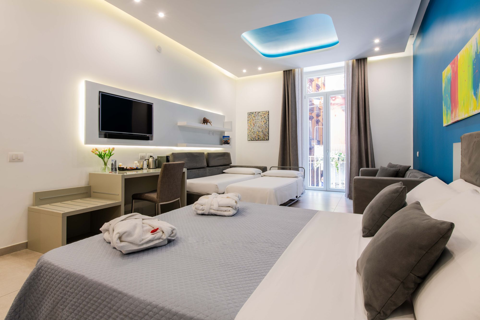 Suite con letto matrimoniale in primo piano, divano letto apribile, parete blu con quadro colorato, TV e balcone - Toledo16 Luxury Suites Napoli