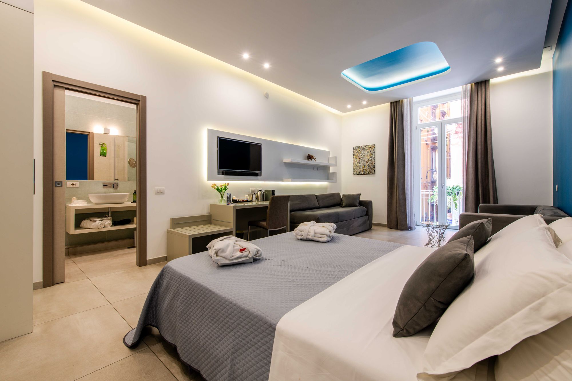 Suite con vista sul bagno dalla camera, TV a parete, scrivania e divano grigio, letto con plaid e accappatoi, porta finestra su balcone - Toledo16 Luxury Suites Napoli