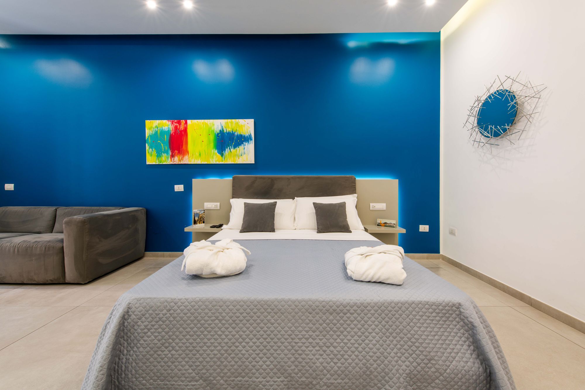Testiera grigia del letto con cuscini bianchi e grigi, quadro astratto multicolore e specchio a raggiera sulla parete bianca - Toledo16 Luxury Suites Napoli