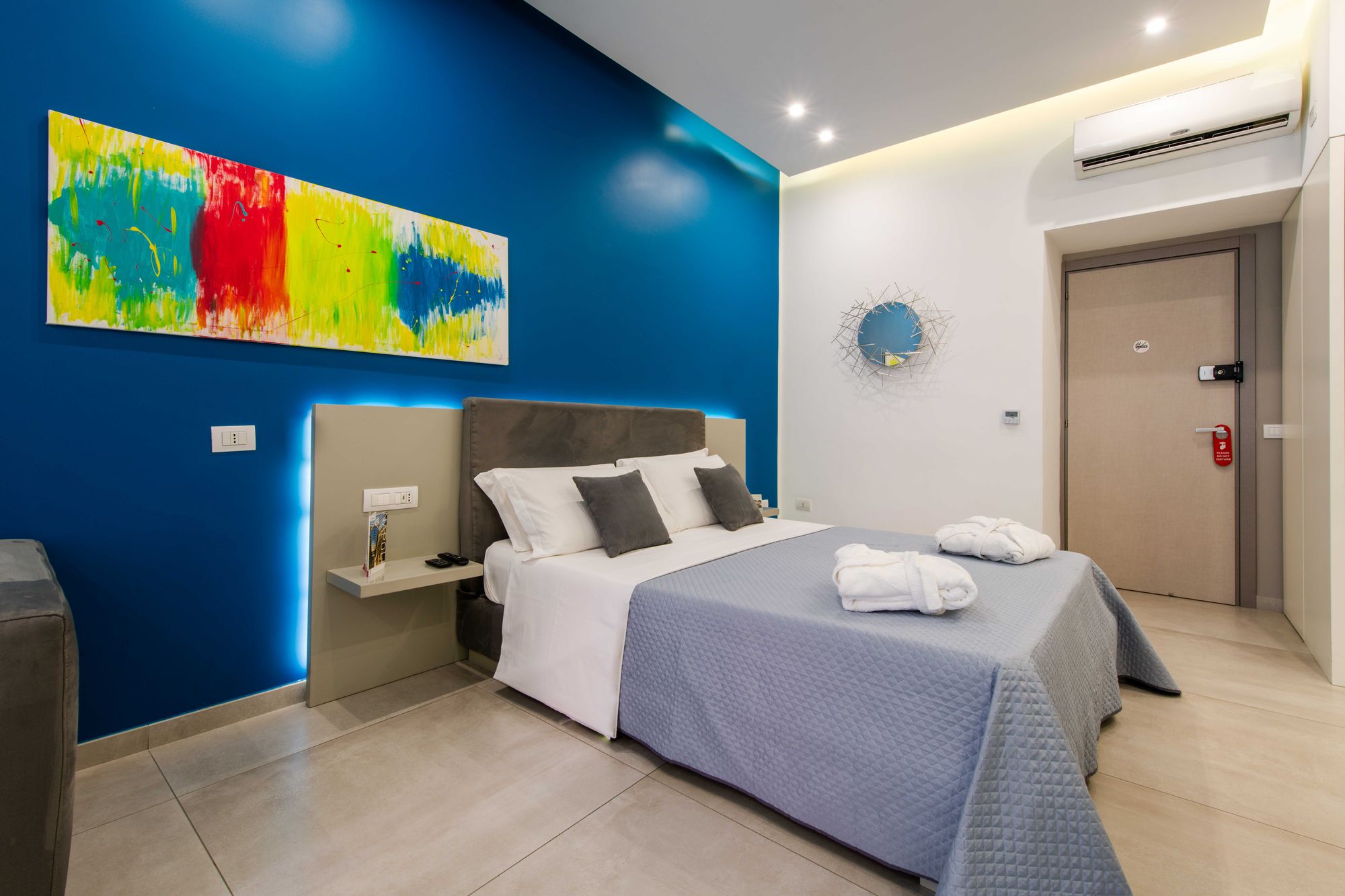 Dettaglio del quadro astratto colorato sulla parete blu, specchio a raggiera e porta d'ingresso con cartello non disturbare - Toledo16 Luxury Suites Napoli