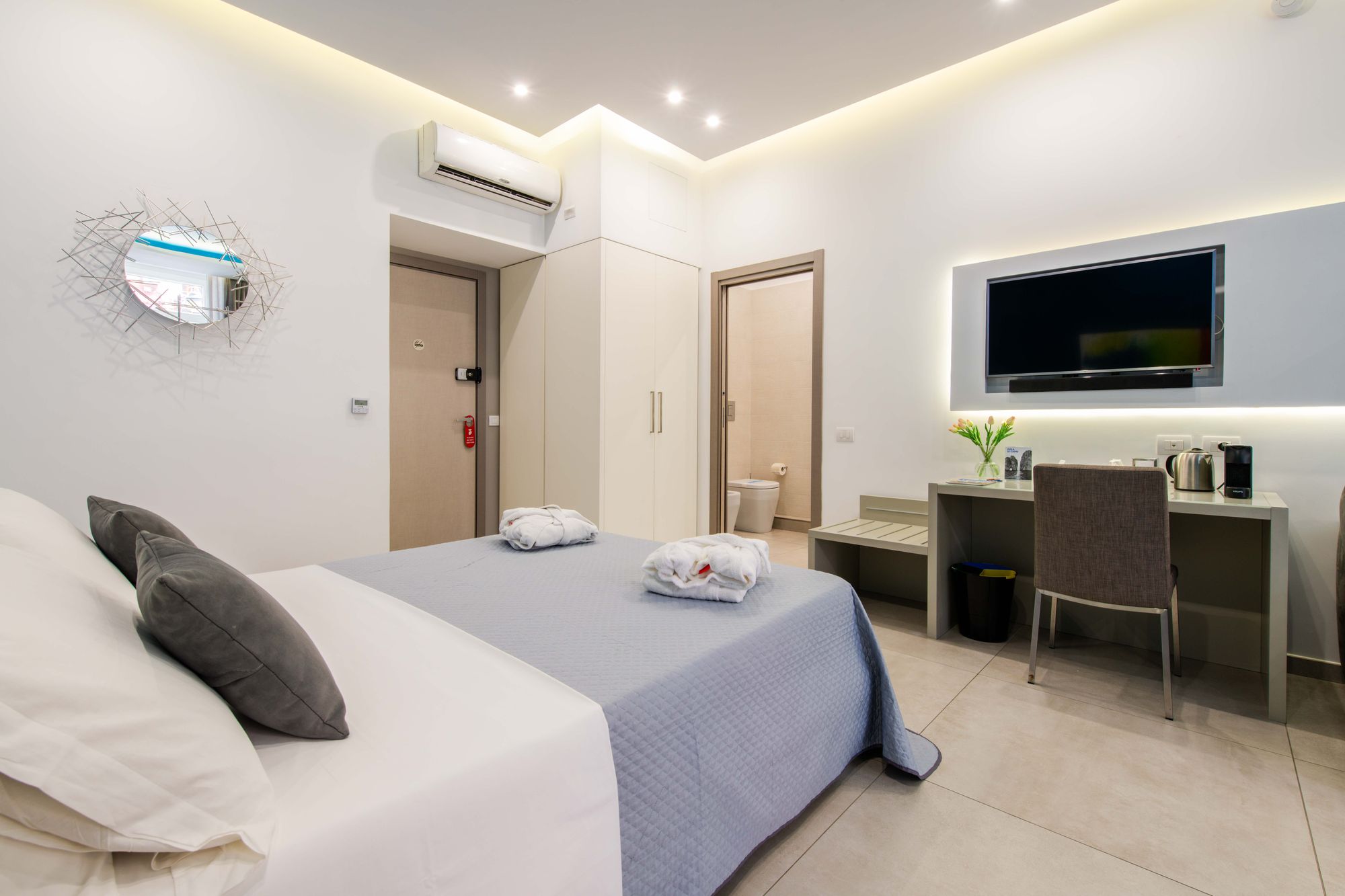 Camera con specchio a raggiera, armadio bianco, porta d'ingresso e scorcio del bagno, TV a parete e scrivania con fiori - Toledo16 Luxury Suites Napoli