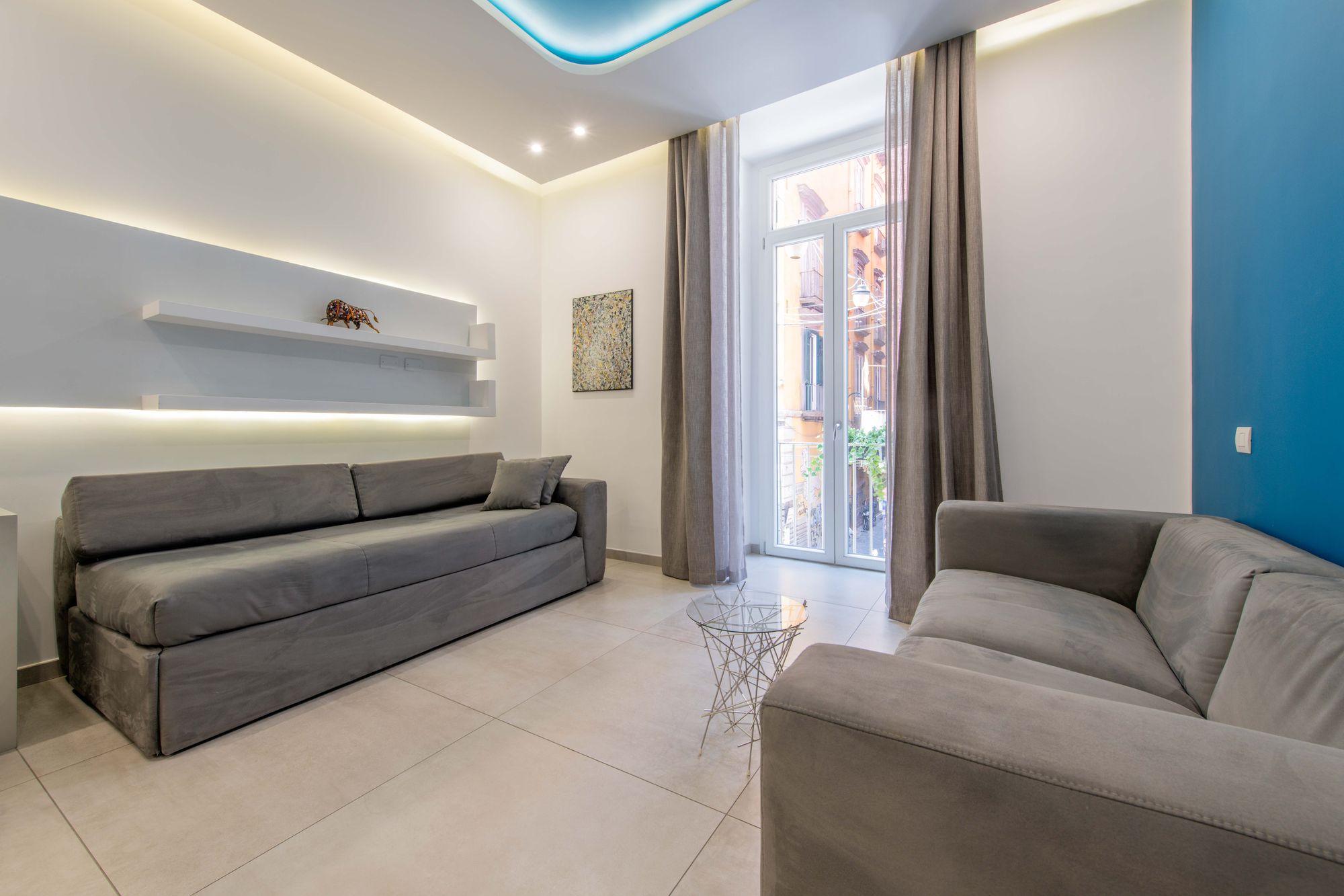 Angolo salotto con divano grigio, mensola con statuetta, tende e porta finestra su balcone affacciata su Via Toledo - Toledo16 Luxury Suites Napoli