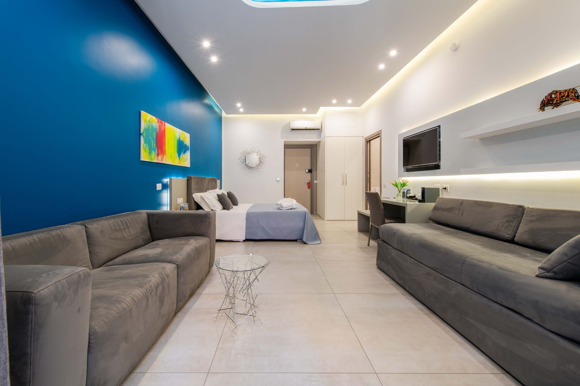 Suite ampia con due divani grigi, letto matrimoniale su parete blu, TV a parete e mensole con illuminazione soffusa - Toledo16 Luxury Suites Napoli