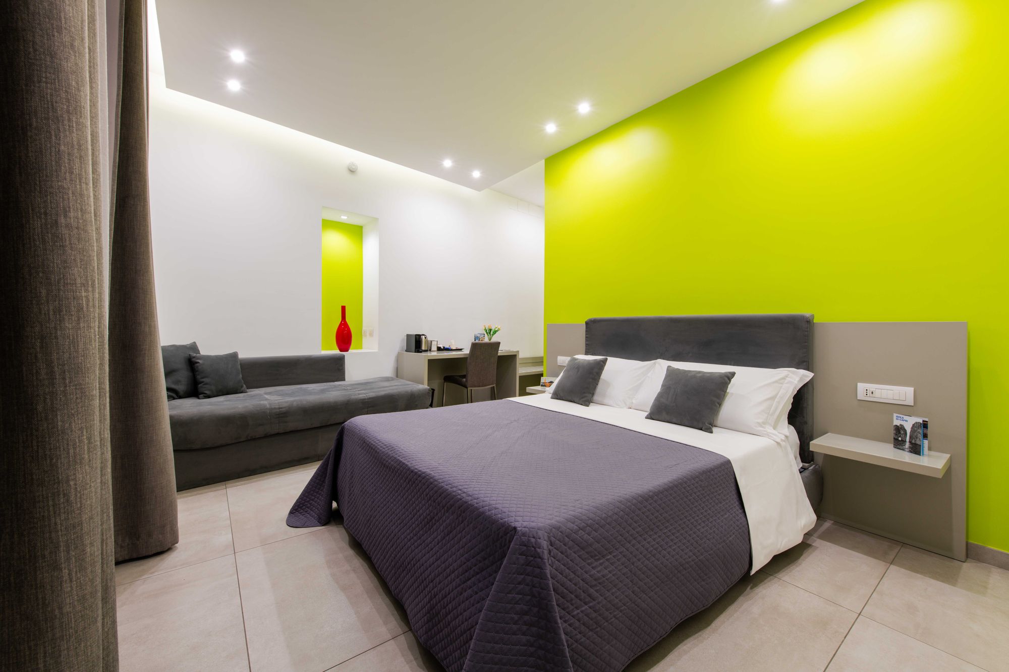 Camera tripla Deluxe con parete verde lime, letto matrimoniale viola, divano grigio e nicchia con vaso rosso decorativo - Toledo16 Luxury Suites Napoli