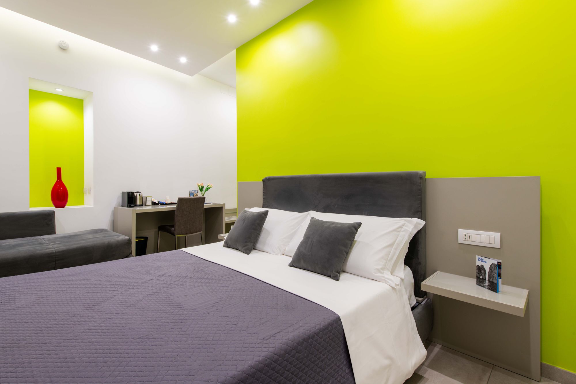 Testata del letto matrimoniale su parete verde lime, comodino sospeso e nicchia con vaso rosso, scrivania sullo sfondo, camera Napoli - Toledo16 Luxury Suites Napoli