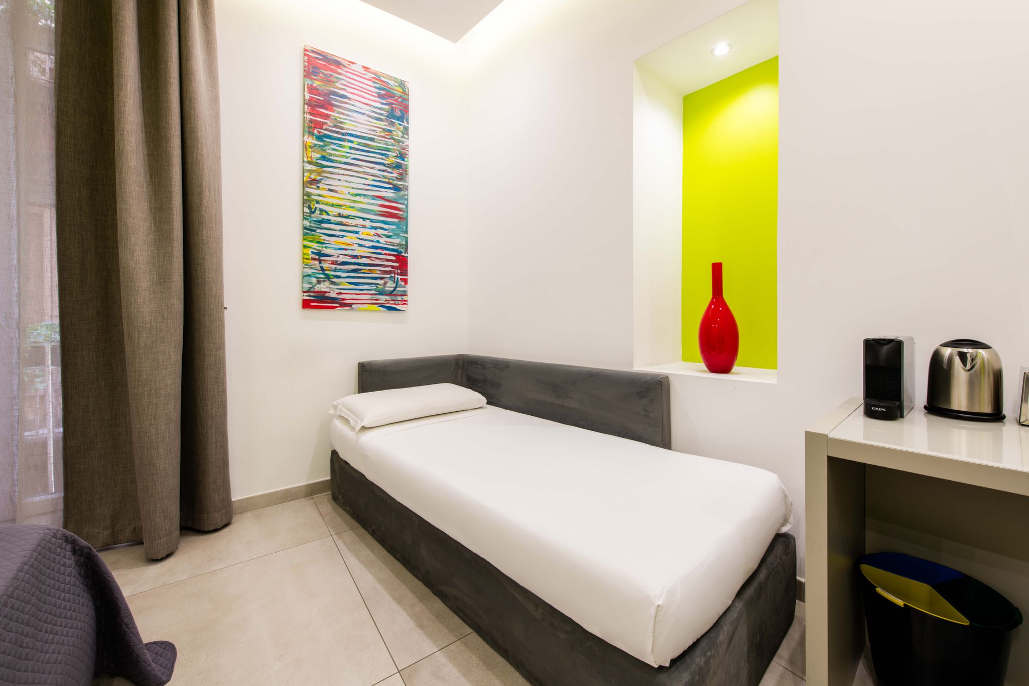 Letto singolo ad angolo con lenzuola bianche, quadro colorato astratto e nicchia con vaso rosso, scrivania con macchina da caffè sullo sfondo, camera Napoli - Toledo16 Luxury Suites Napoli