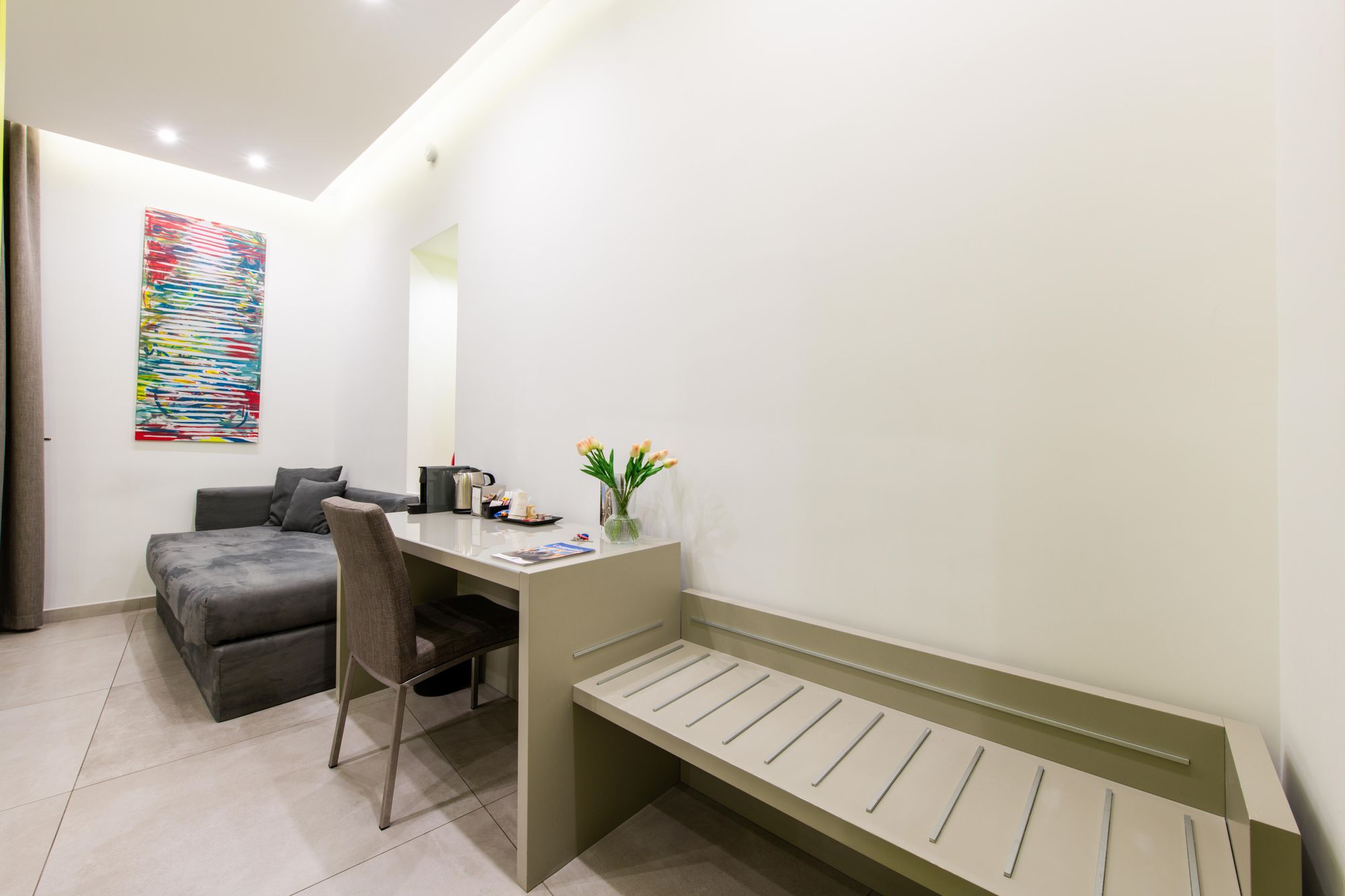 Angolo soggiorno con divano grigio in velluto, scrivania, panca imbottita e quadro astratto colorato, camera Napoli - Toledo16 Luxury Suites Napoli