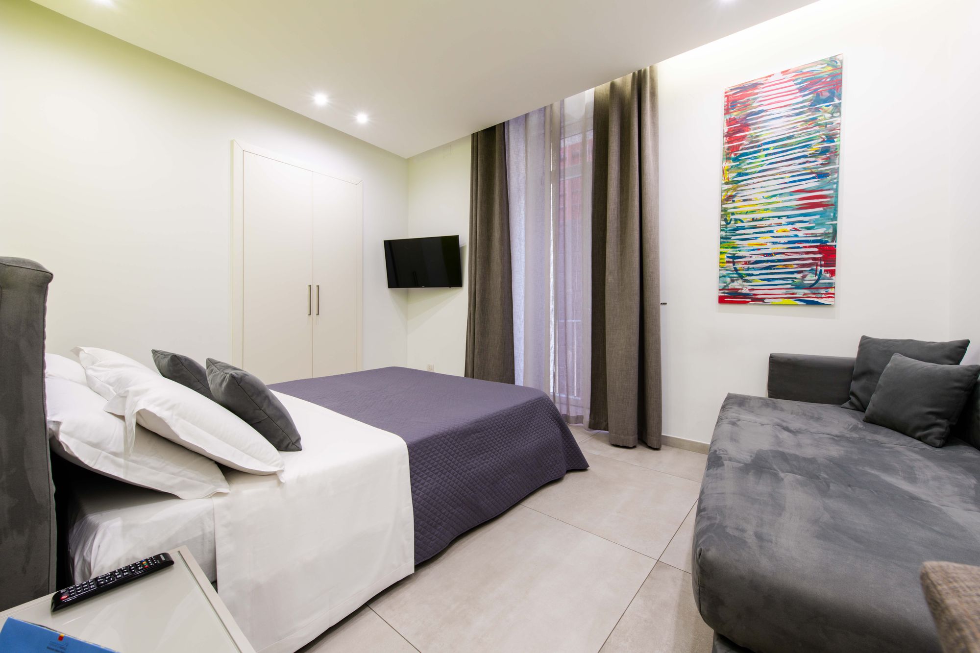 Letto matrimoniale con copriletto viola scuro, testiera grigia, armadio bianco e TV a parete, tende lunghe sulla porta finestra del balcone affacciato su Via Toledo, camera Napoli centro - Toledo16 Luxury Suites Napoli