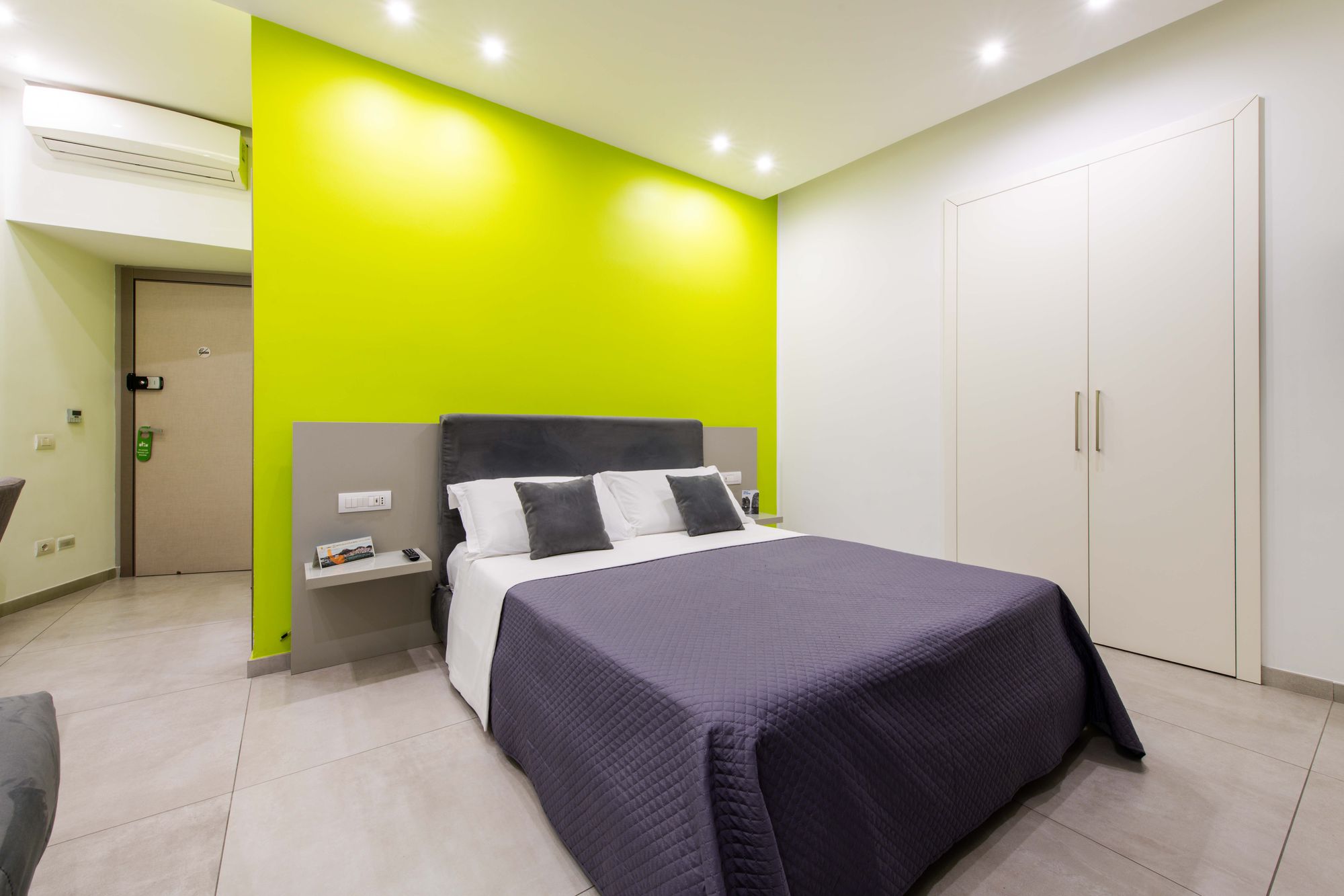 Letto matrimoniale con copriletto viola scuro davanti a parete verde lime, comodini sospesi e armadio bianco sullo sfondo, camera Napoli - Toledo16 Luxury Suites Napoli