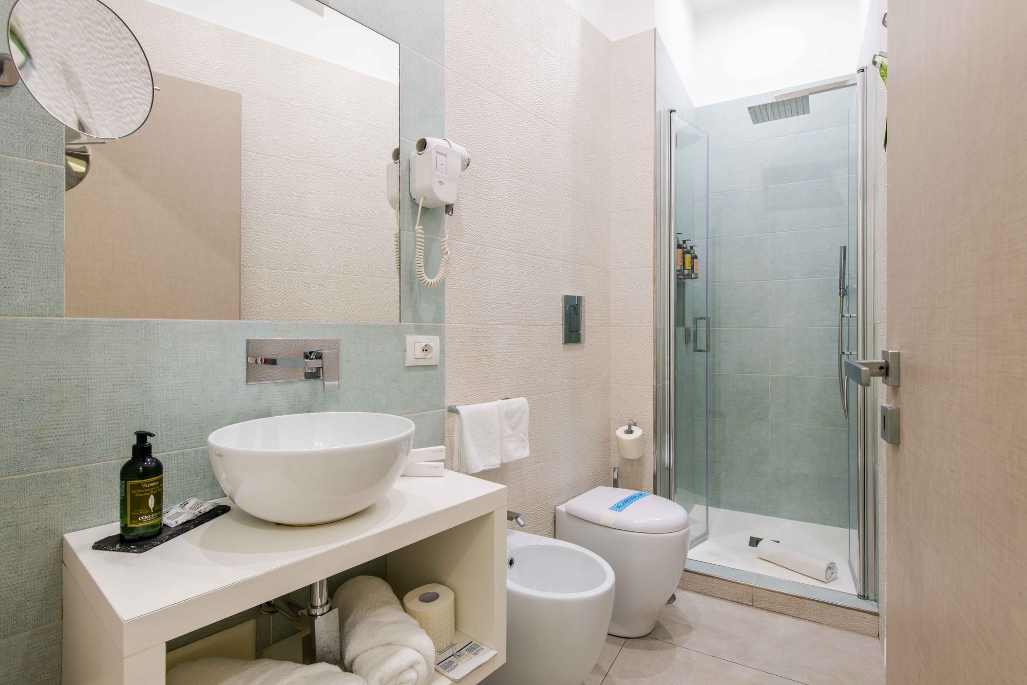 Bagno con lavabo a catino bianco su piano beige, specchio con asciugacapelli e box doccia in vetro con sanitari - Toledo16 Luxury Suites Napoli