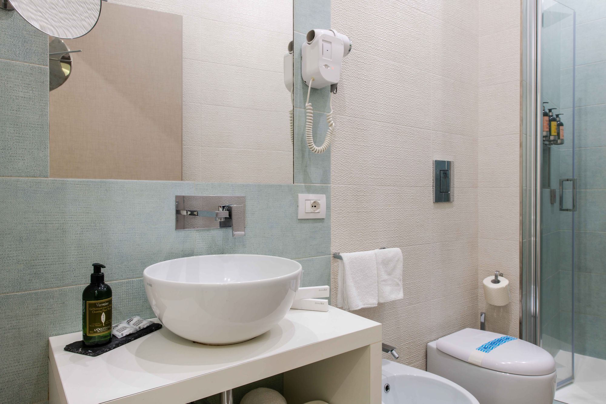 Lavabo a catino bianco con rubinetteria a parete cromata, asciugacapelli e box doccia in vetro sullo sfondo - Toledo16 Luxury Suites Napoli
