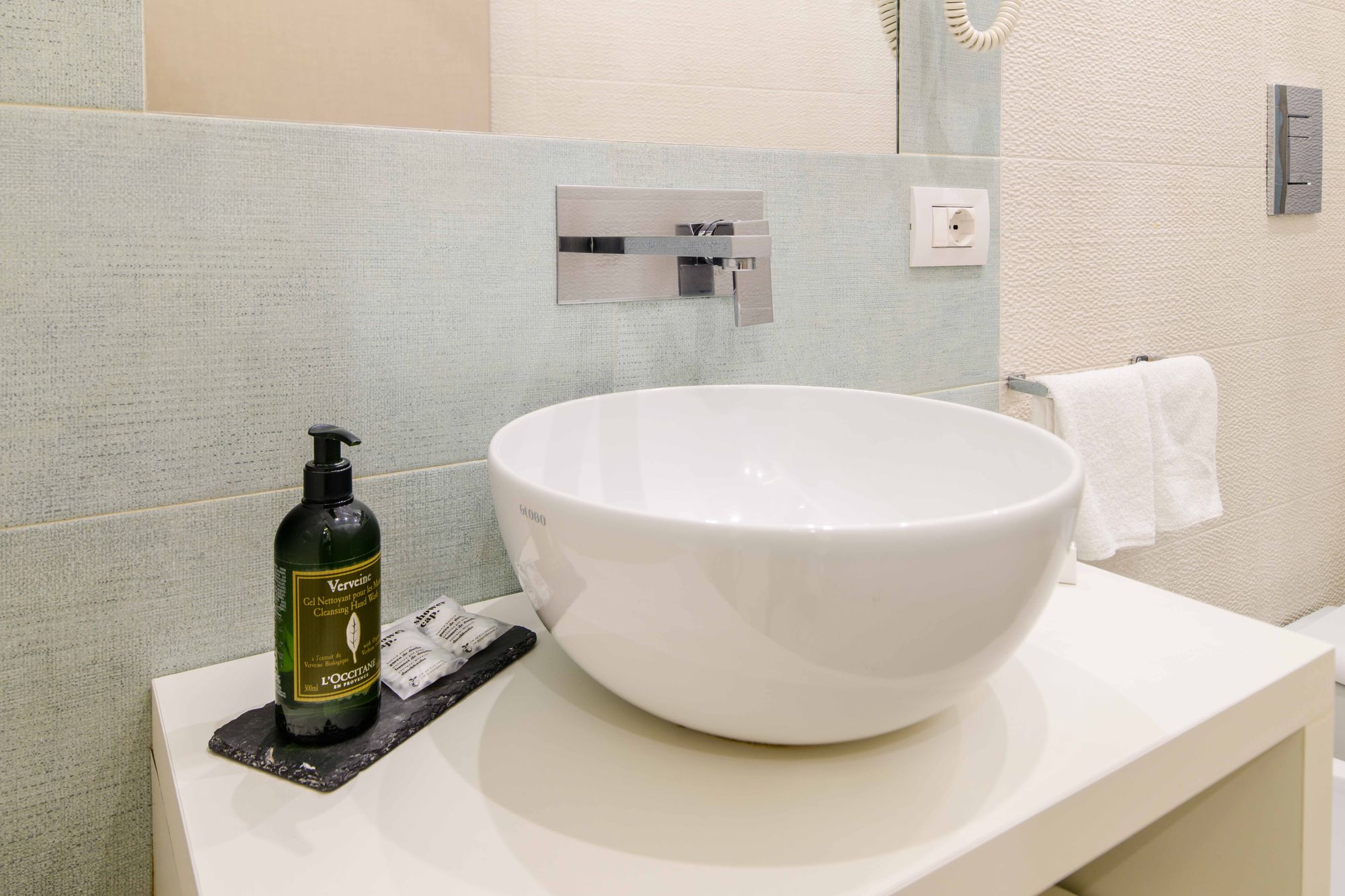 Dettaglio del lavabo a catino con rubinetto a parete, flacone di sapone L'Occitane e asciugamano bianco, bagno camera Napoli - Toledo16 Luxury Suites Napoli