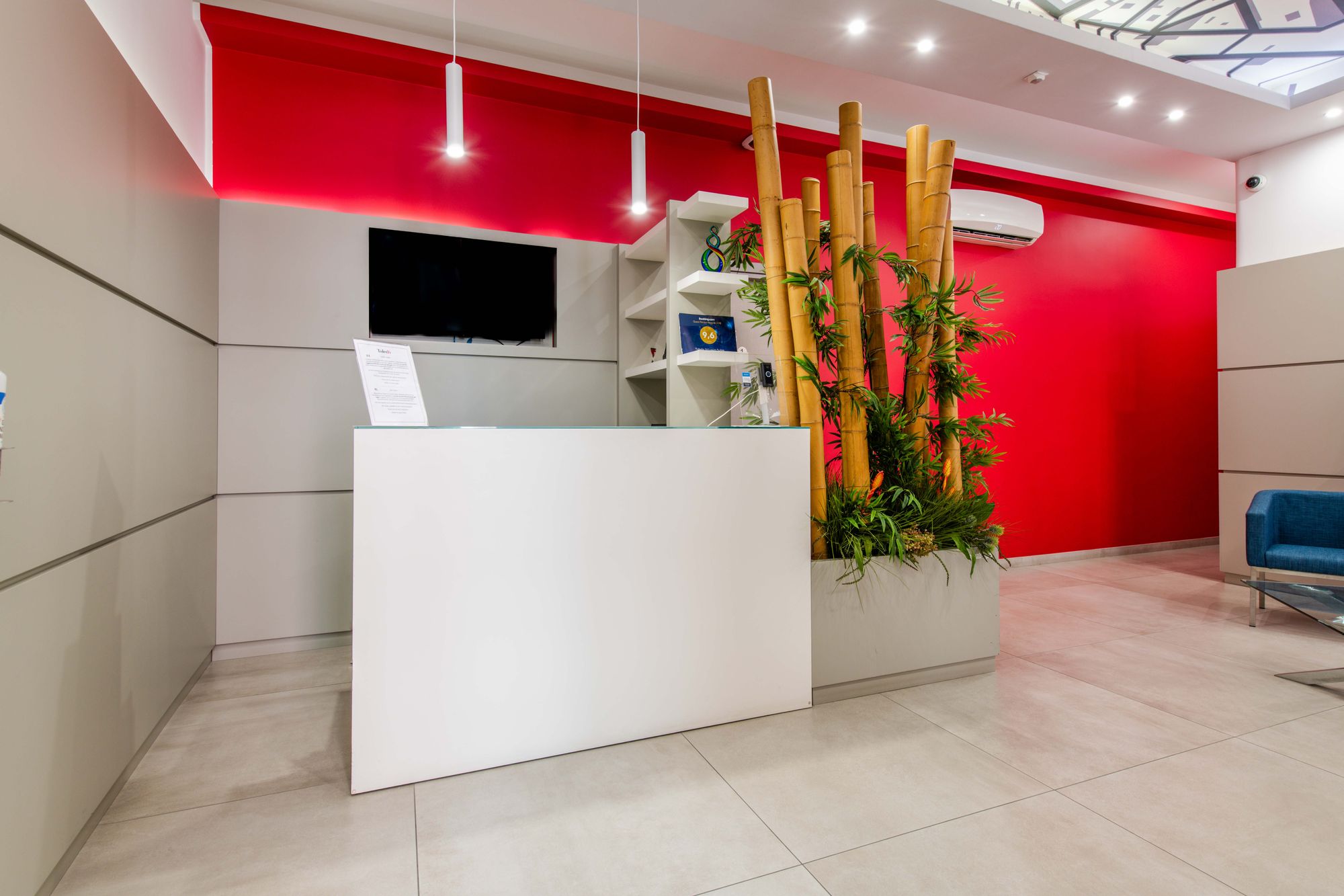 Banco reception bianco con parete rossa, bambù decorativo e televisore a parete, Toledo16 Luxury Suites Napoli centro