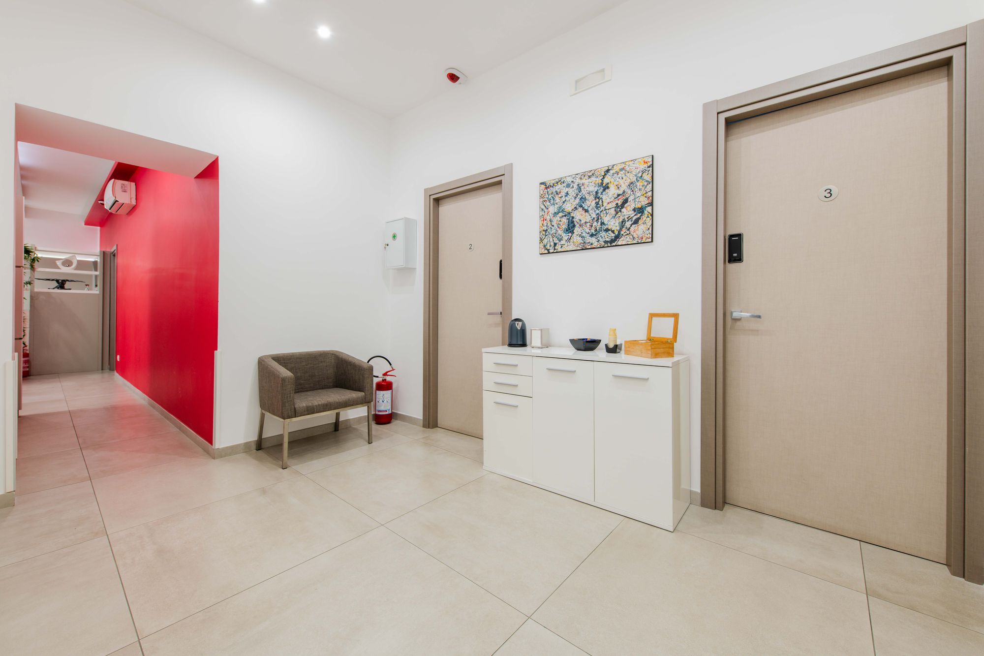 Corridoio con parete rossa sullo sfondo, porte delle camere e poltrona grigia, Toledo16 Luxury Suites Napoli centro